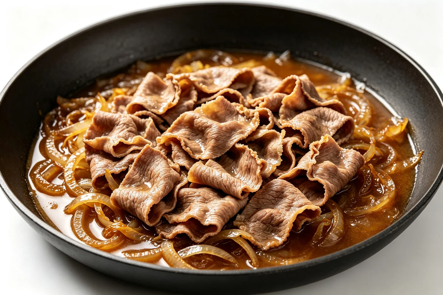 Paper-thin gyudon beef ribbons and fully softened golden onions simmering in glossy amber soy-dashi broth in a wide dark pan on white surface