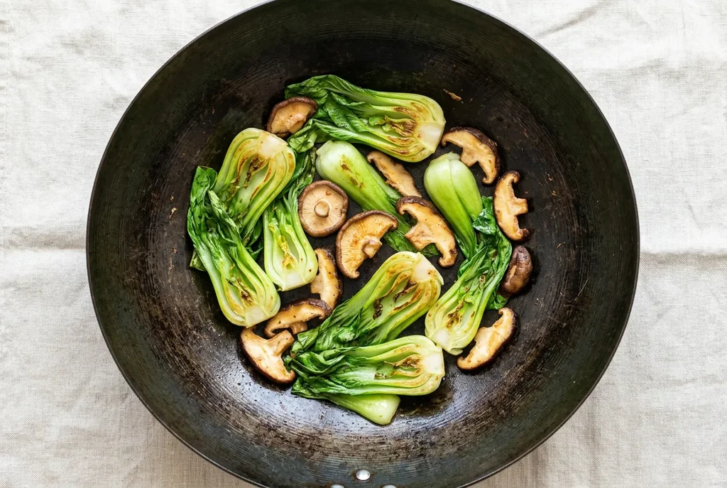  Baby bok choy halves and sliced shiitake mushrooms mid stir-fry in a carbon steel wok showing caramelised edges on linen surface