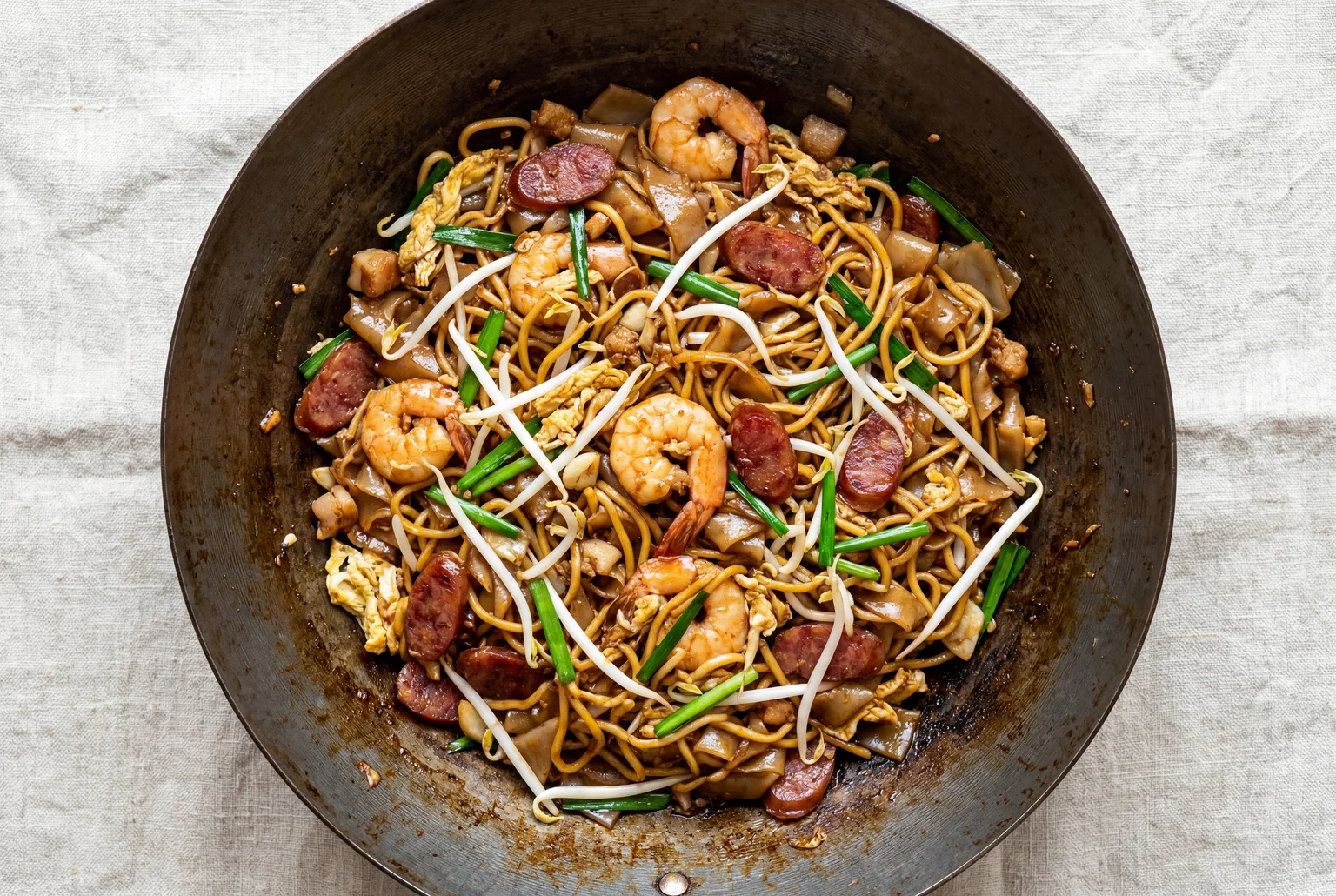 Char Kway Teow noodles mid stir-fry in a carbon steel wok — flat rice noodles and yellow noodles coated in dark soy glaze with prawns, lap cheong and egg ribbons on linen surface