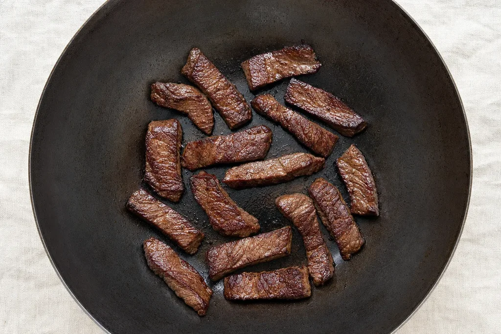 Velveted flank steak strips searing in a single layer in a carbon steel wok showing deep mahogany crust on linen surface