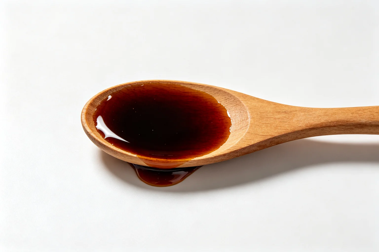 Wooden spoon with dark amber teriyaki glaze pooling inside and dripping onto a white surface showing the thick glossy consistency of a properly reduced hon-mirin glaze
