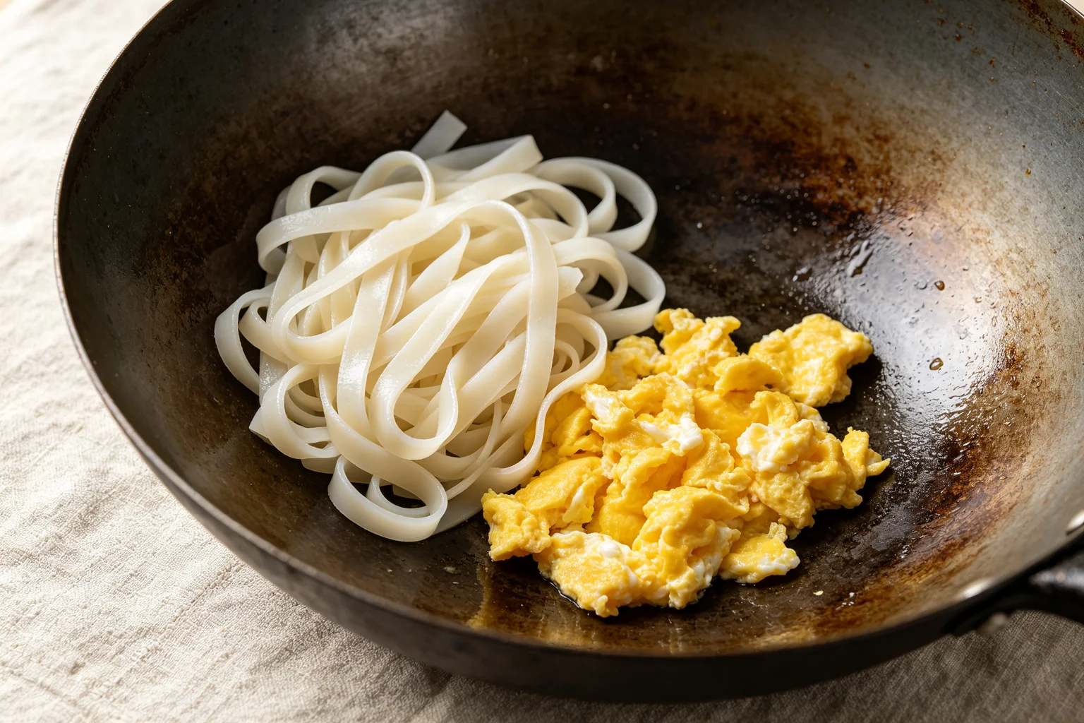  Beaten egg scrambling in the empty section of a dark wok beside pad thai noodles pushed to one side showing the separate egg technique on linen surface