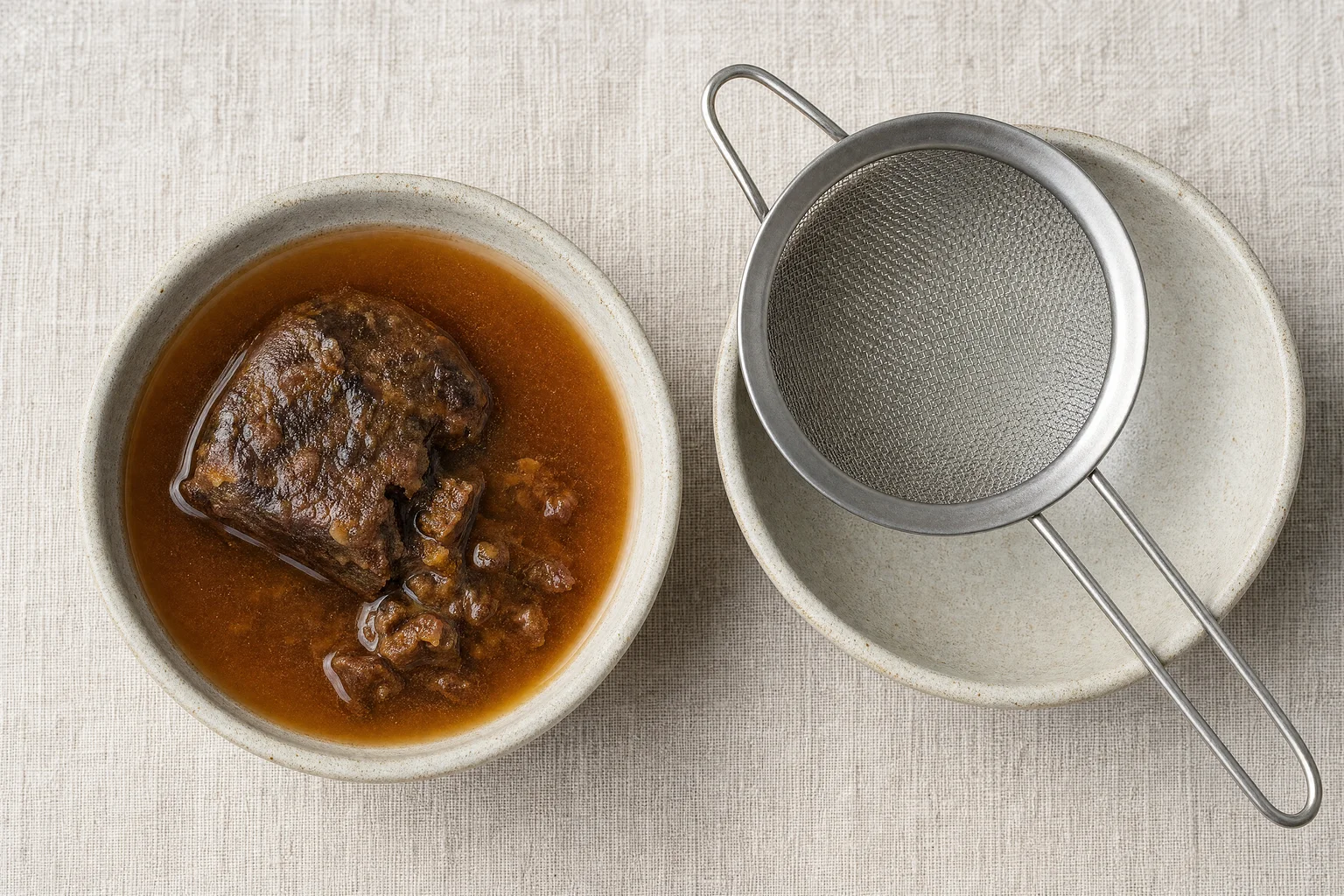 Tamarind block soaking in warm water in a ceramic bowl turning amber-brown, with a fine mesh strainer ready alongside on linen surface