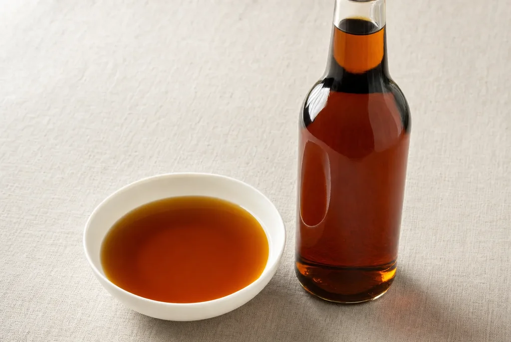 Small fish sauce bottle and ceramic bowl of amber liquid next to a bowl of golden broth on linen — showing fish sauce as a finishing ingredient